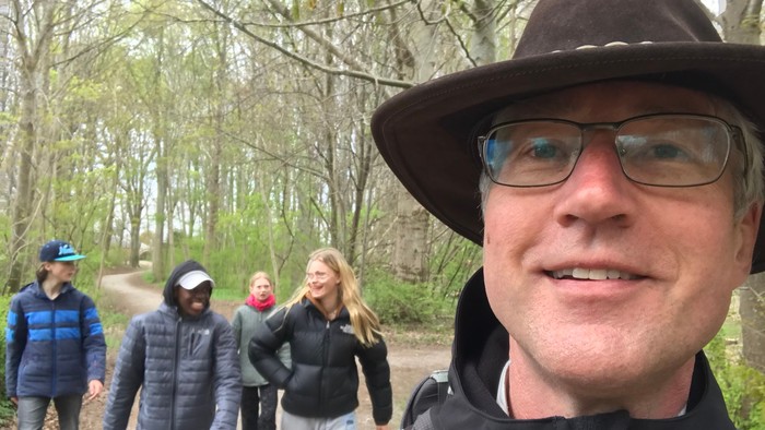 En mand i hat og briller tar en selvie med en gruppe på en skovsti