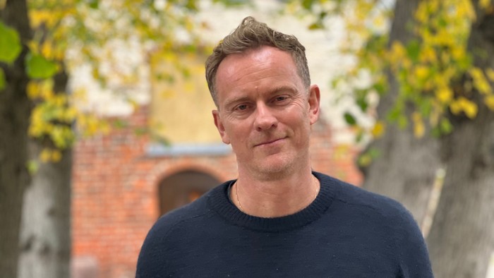 En mand står i forgrunden med et smil på et træ og et murværk bagved.