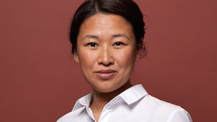 En kvinde med mørke hår og et hvidt blus, står foran en rød mur.