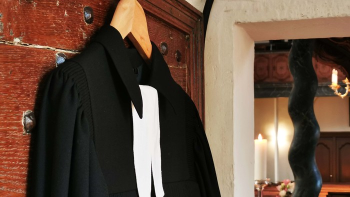 Ein schwarzer Talar mit weißer Schleife hängt an einem Kleiderbügel an einer roten Ziegelwand.