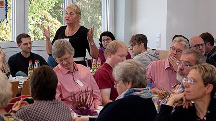 Eine Gruppe von Menschen sitzt an Tischen und hört einer Frau zu, die aufsteht und spricht.