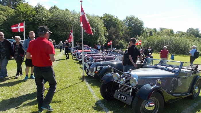 En gruppe mennesker og gamle biler på en græsplads med danske flag.