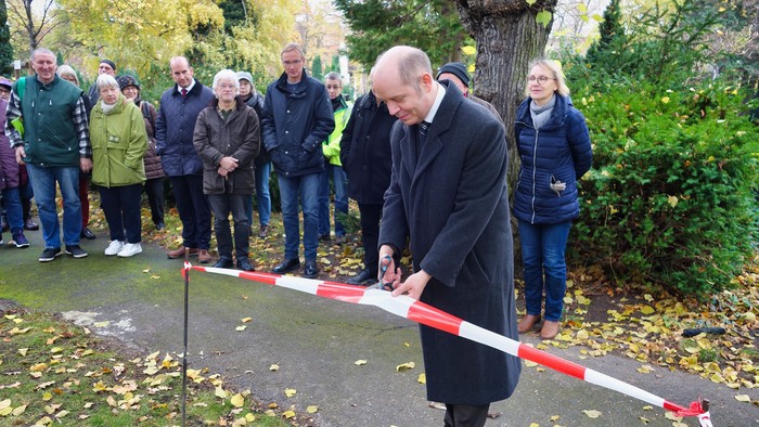 Ein Mann in einem Gewand schneidet ein Band durch, während eine aufmerksame Menge in einem Park im Freien zusieht.