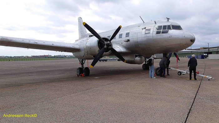 Großes silbernes Propellerflugzeug auf Rollfeld mit mehreren Menschen.