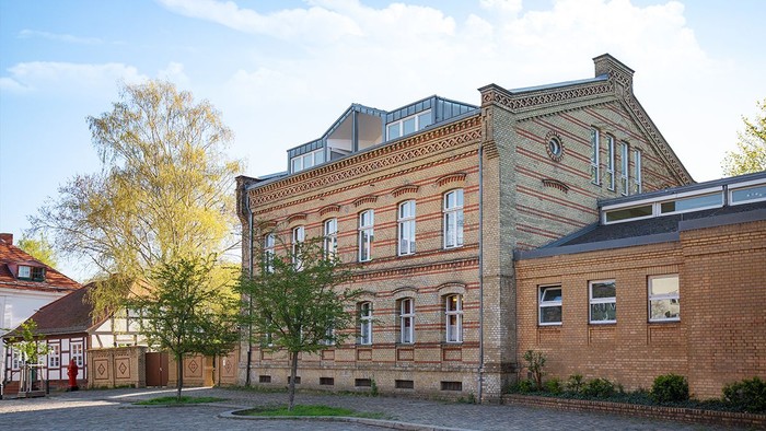 Historisches Gebäude mit Ziegelfassade und großen Fenstern, umgeben von Bäumen und Kopfsteinpflasterstraße