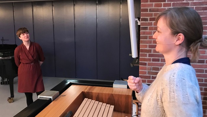 To da kvinder står i et rum med mørke vægge og røde mursten. En kvinde holder en lysestage.