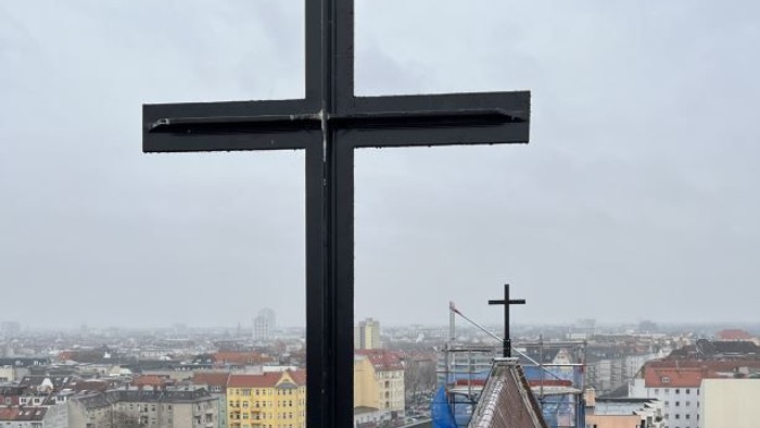 Auffälliges schwarzes Kreuz auf einem Gebäude mit Blick über eine Stadtlandschaft mit Bahngleisen und urbanen Strukturen.
