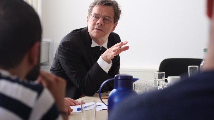 Mann in Anzug spricht gestikulierend mit anderen am Tisch.