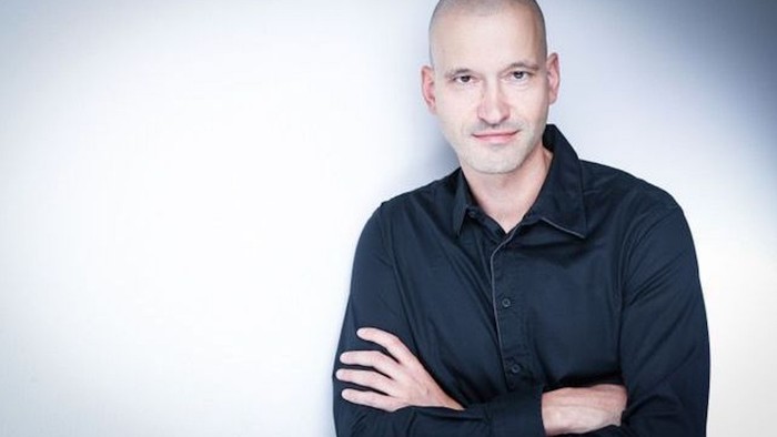 Ein kahlköpfiger Mann mit verschränkten Armen in einem dunklen Hemd.