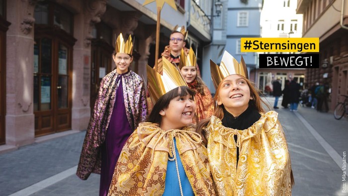 Eine Gruppe von Menschen in prächtigen, goldenen Gewändern und Kronen feiert auf einer Straße, mit einem Banner 