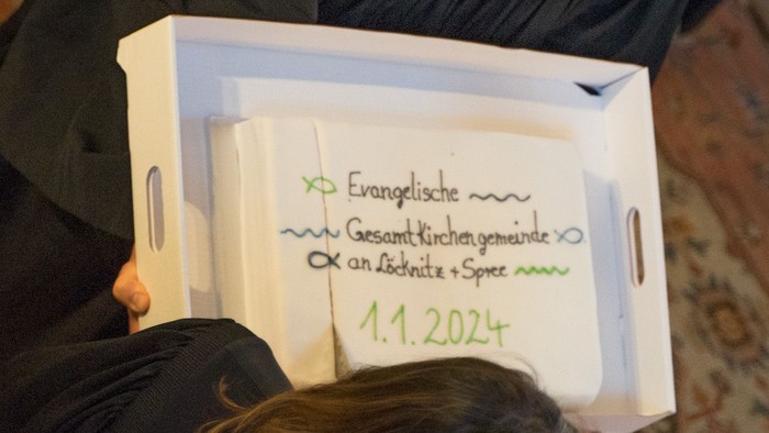 Ein Mann in schwarzer Robe hält eine Kiste mit Aufschriften, während eine Frau in schwarzem Kleid daneben steht.