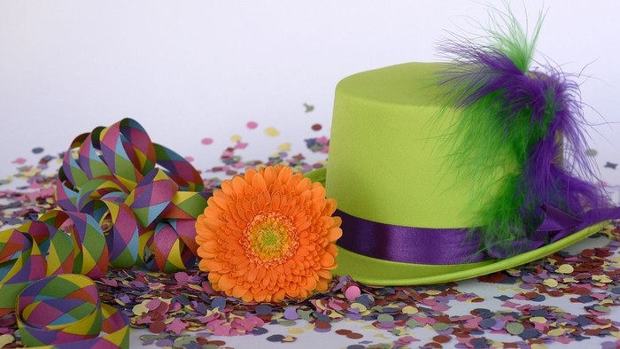 Ein grüner Zylinder mit lila Feder, eine orange Blume und bunte Papierschnitzel auf weißem Hintergrund