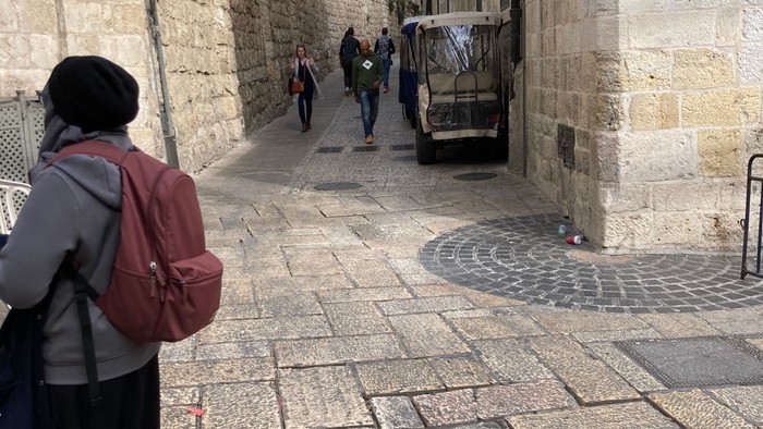 Frau mit Rucksack geht auf Kopfsteinpflasterstraße, Menschen und Bus im Hintergrund