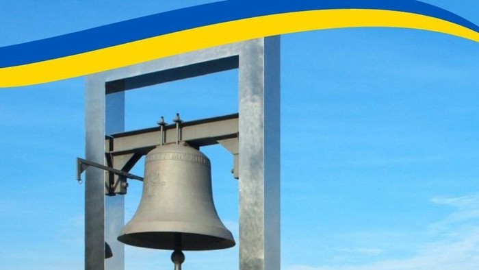 Eine Glocke hängt an einem Metallrahmen vor blauem Himmel mit blau-gelber Flagge.