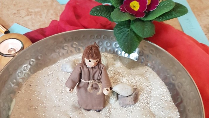 In der silbernen Schale sitzt eine kleine Figur im Schnee, umgeben von Steinen und einer Blume.