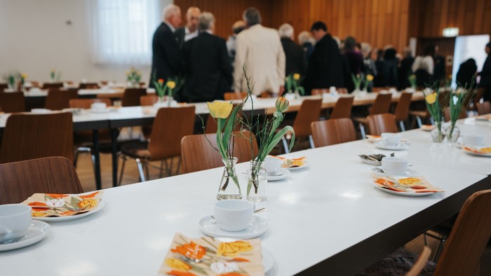 Ein langer Tisch mit Blumen, Tassen und Tellern, umgeben von Stühlen, in einem Raum mit Menschen im Hintergrund
