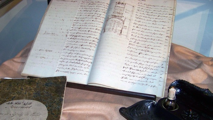 Ein offenes Buch mit handgeschriebenem Text und ein kleines Buch liegen auf einem Tisch.