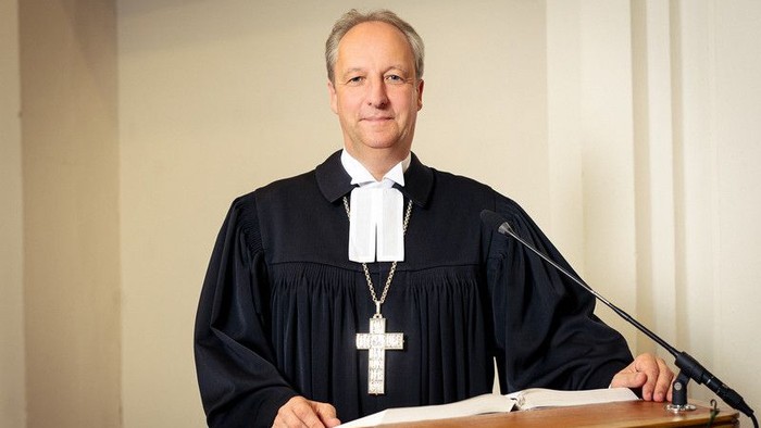Richter in schwarzer Robe, der an einem Rednerpult mit einem Kreuzanhänger und Papieren spricht.