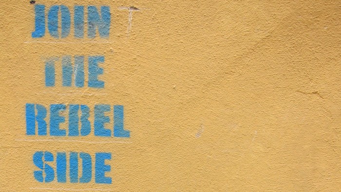 Blauer Graffiti-Text auf gelber Wand: "JOIN THE REBEL SIDE"