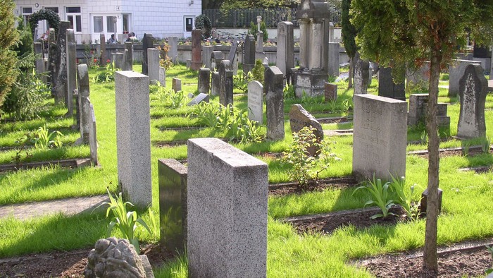 Ein Friedhof mit vielen Grabsteinen und Bäumen, im Hintergrund ein weißes Gebäude.