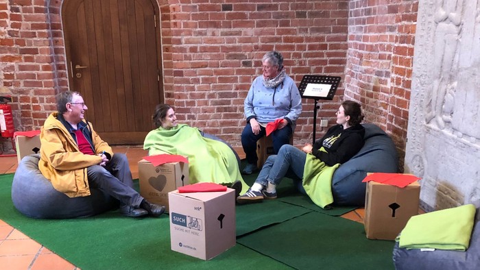 Vier Personen sitzen auf Kissen und Kartons vor einer Ziegelwand.