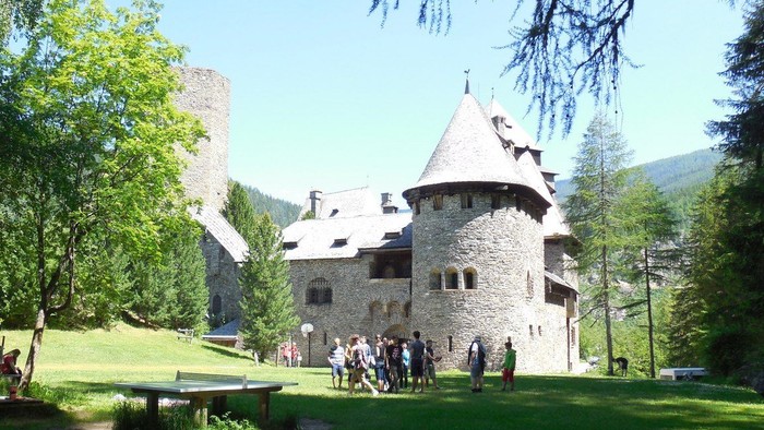 Ein Steinhaus mit Turm, umgeben von Bäumen und Menschen auf der Wiese