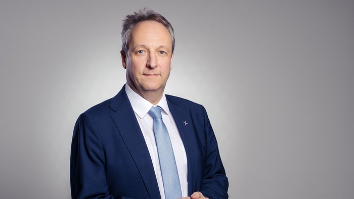 Professioneller Mann in dunklem Anzug und hellblauer Krawatte, der selbstbewusst vor einem neutralen Hintergrund steht.