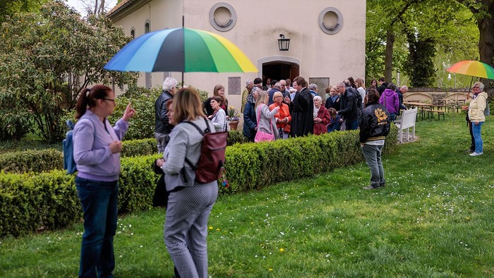 Eine Gruppe von Menschen steht auf einem grasbewachsenen Bereich vor einem Gebäude, einige halten bunte Regenschirme.