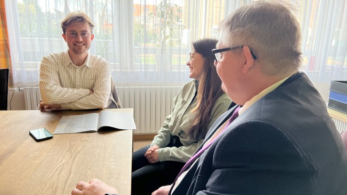 Drei Personen sitzen an einem Tisch in einem Büro, eine Person lächelt.