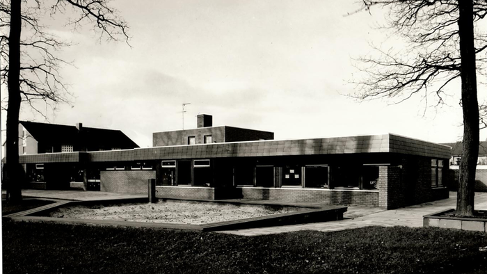 Altes Gebäude mit flachem Dach und großen Fenstern, umgeben von kahlen Bäumen und einem gepflegten Rasen.