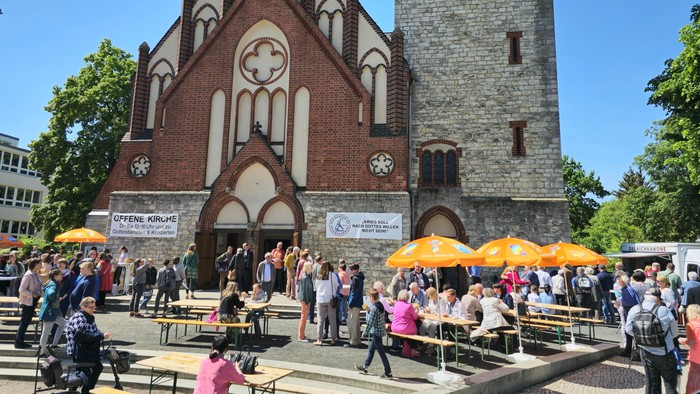 Vor einer Kirche sitzen Menschen unter gelben Schirmen im Freien.