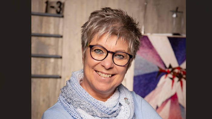 Eine Frau mit kurzem Haar und Brille lächelt vor einem bunten Hintergrund.