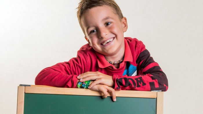 Ein fröhliches Kind lehnt an einer grünen Tafel und lächelt breit.