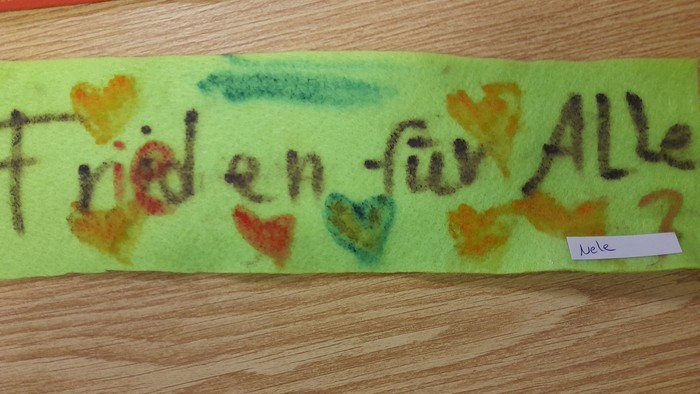 Grüner Filzbanner mit bunten Handabdrücken und dem Text *„Frieden für Alle“*