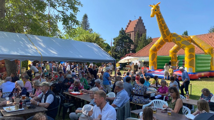 En folkelig fest med mange mennesker, der sidder ved bord og gennyder mad og drikke. Der er en stor gallaumgang med et giraffemotiv og en blå telt.