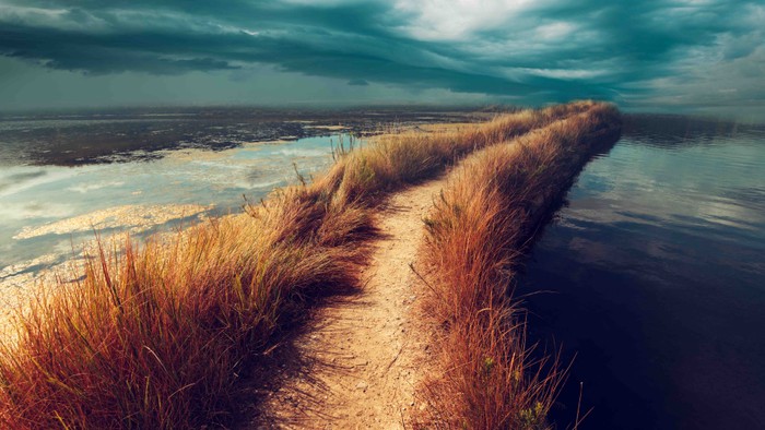 Ein schmaler Pfad führt durch trockenes Gras zur Wasserfläche unter einem bewölkten Himmel