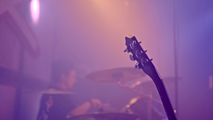 Blick auf eine vernebelte Bühne, getaucht in buntes Licht. Im Vordergrund ist der Kopf einer Gitarre zu sehen, im Hintergrund ein Schlagzeug.