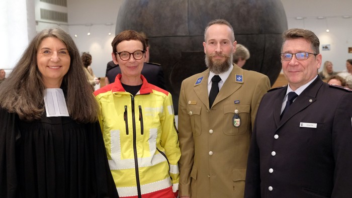 Vier Personen stehen vor einer großen Kugel, zwei in Uniformen, eine in einem gelben Sicherheitsoverall und eine in einem schwarzen Kleid.