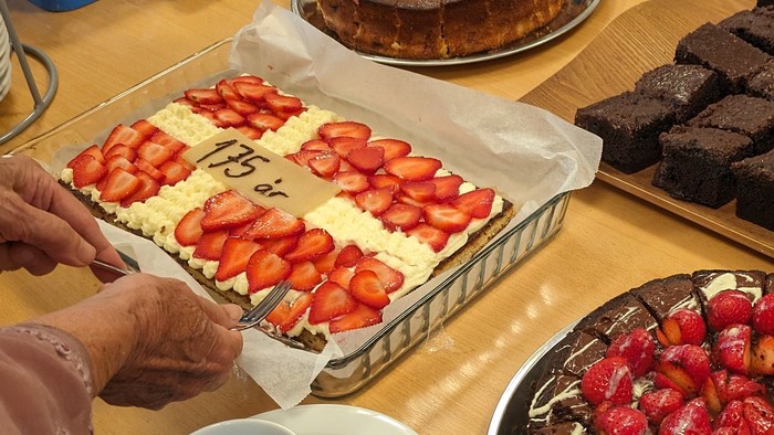 En person serverer kage på et bord med forskellige kager og drikkevarer