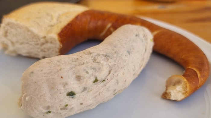 Auf dem Teller liegen eine Wurst und ein Brötchen, daneben ein Glas.