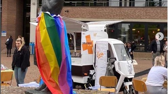 Statue mit Regenbogenfahne vor Gebäude mit Menschen und Fahrzeugen