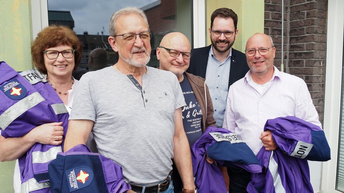 Eine Gruppe von fünf Personen hält lila und weiße Trikots und posiert vor einem Haus.