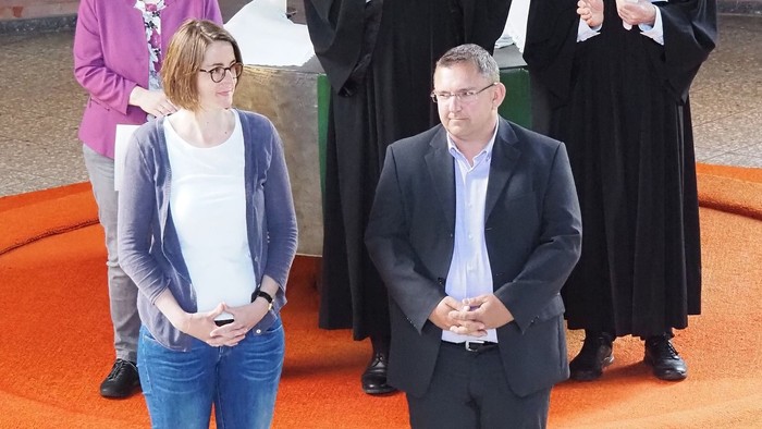 Zwei Priester in schwarzen Gewändern stehen hinter einem Mann und einer Frau, die vor ihnen stehen.