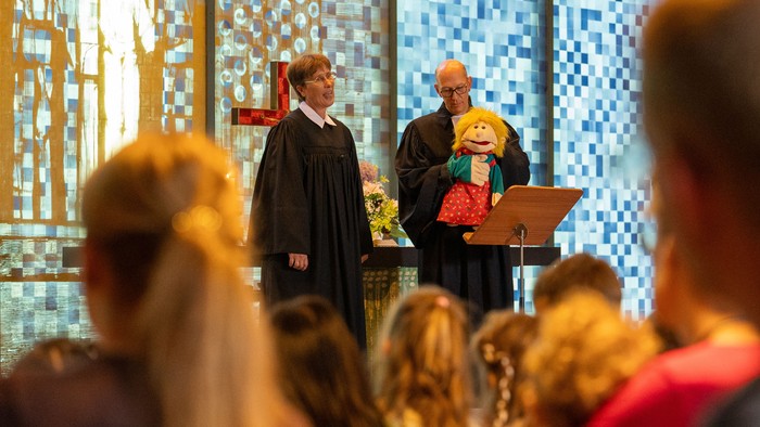 Eine Frau in schwarzer Robe steht vor einem Podium mit Mikrofon, während eine Person in einem bunten Outfit daneben steht. Im Hintergrund befindet sich ein blaues Mosaikfenster.