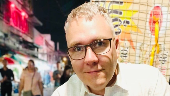 En ung mand med briller og blond hår står i en lebendig bygate med farverige lys og andre mennesker i baggrunden.