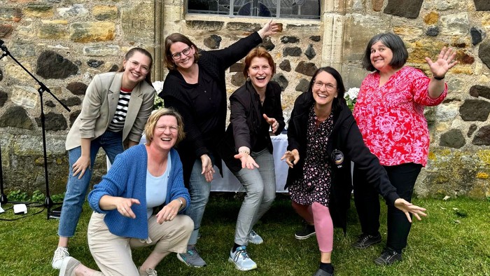 Gruppe von sechs Personen, die vor einem Steingebäude mit Rundbogenfenster posieren.
