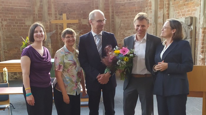 Eine Gruppe von fünf Personen posiert in einem Kirchenraum, eine hält einen Blumenstrauß