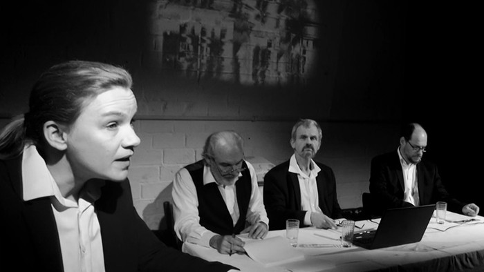 Eine Gruppe von Menschen sitzt an einem Tisch und diskutiert, im Hintergrund ist ein Projektionsbild zu sehen.