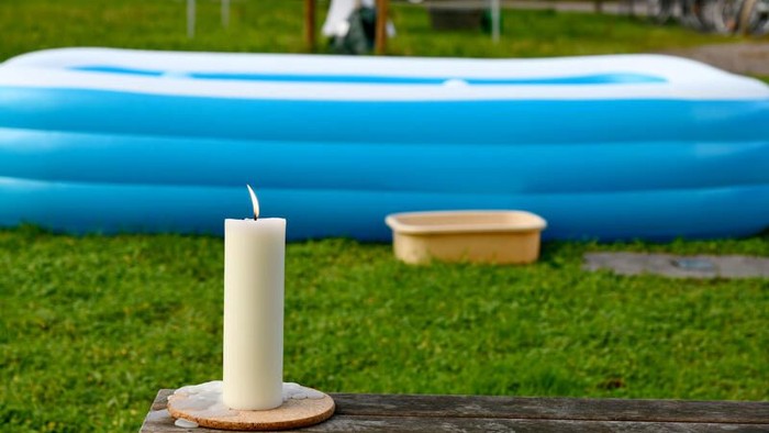 Im Vordergrund eine brennende Kerze auf einem Holztisch, im Hintergrund ein aufgeblasenes blaues Becken auf dem Rasen.