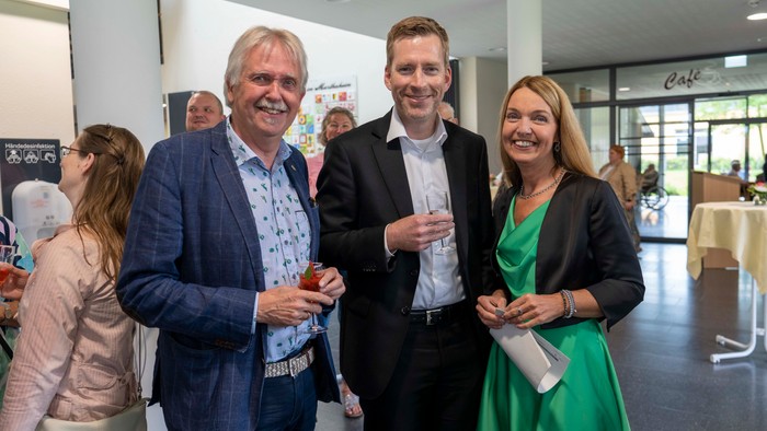 Drei Personen lächeln in einem Raum, eine Frau in Grün hält eine Mappe